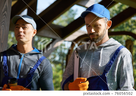 Portrait of two confident young repairmen in uniform standing outdoors with papers while working on construction project Portrait of two confident young repairmen in uniform standing outdoors with papers while working on construction project 76056502