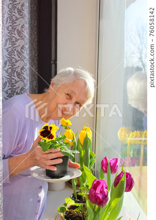 Old lonely woman sitting near the window in his house. 76058142