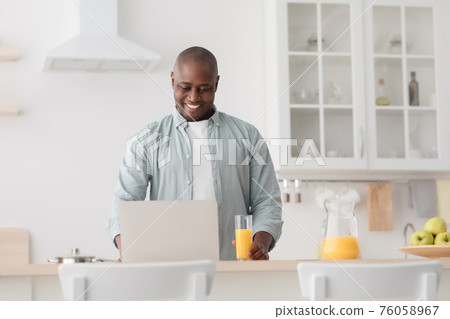 Happy african american mature man drinking orange juice and watching movie on laptop in kitchen Happy african american mature man drinking orange juice and watching movie on laptop in kitchen 76058967