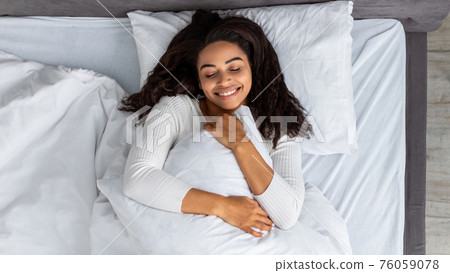 Satisfied young black woman waking up and hugging pillow Satisfied young black woman waking up and hugging pillow 76059078