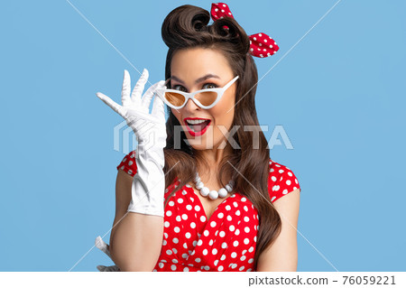Portrait of excited pin up woman in polka dot dress, headband and gloves touching her sunglasses on blue background Portrait of excited pin up woman in polka dot dress, headband and gloves touching her sunglasses on blue background 76059221
