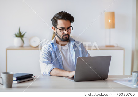 Telecommuting. Eastern Man Using Laptop Computer, Sitting At Desk In Home Office Telecommuting. Eastern Man Using Laptop Computer, Sitting At Desk In Home Office 76059709