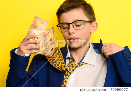 A child with a pathetic look holds in his hands euro bills, a boy in an adult suit on a yellow background, a blue jacket and a yellow tie. A child with a pathetic look holds in his hands euro bills, a boy in an adult suit on a yellow background, a blue jacket and a yellow tie. 76060077