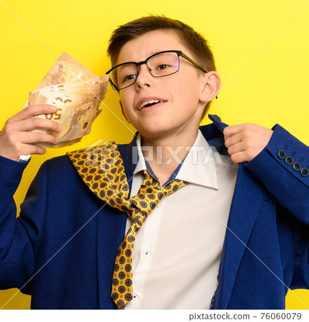 A child with a pathetic look holds in his hands euro bills, a boy in an adult suit on a yellow background, a blue jacket and a yellow tie. A child with a pathetic look holds in his hands euro bills, a boy in an adult suit on a yellow background, a blue jacket and a yellow tie. 76060079