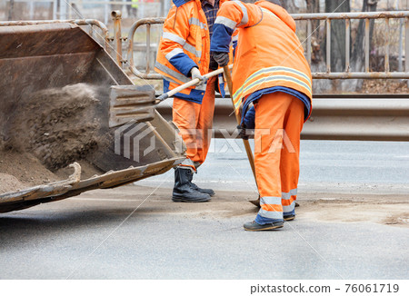 Road workers scrape off accumulated sand between lanes of the road with shovels and load it into a metal grader bucket. 76061719
