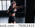 Athletic fit female boxer exercising punches with boxing bag in gym during kickboxing and self defense intensive workout 76063419