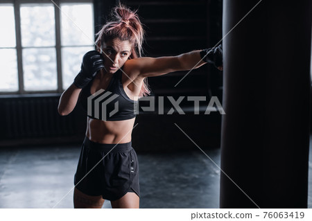 Athletic fit female boxer exercising punches with boxing bag in gym during kickboxing and self defense intensive workout 76063419