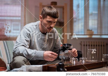A man learns to conduct a tea ceremony at home on his own. Pours black ripe tea with clip into a teapot on a bamboo gongfu tray. A man learns to conduct a tea ceremony at home on his own. Pours black ripe tea with clip into a teapot on a bamboo gongfu tray. 76070380