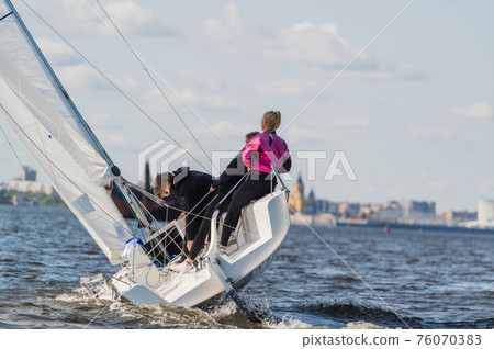 A beautiful picture with a sports yacht on the background of an urban landscape floating under full sails on a wide river. 76070383