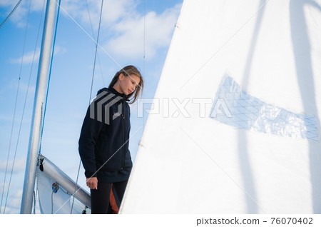 A young blonde athlete on a yacht against a blue sky, a mast and a white sail. 76070402