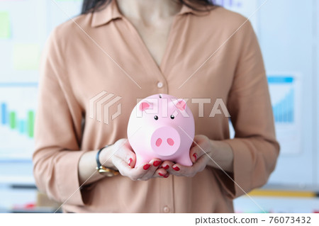 Woman holding pink piggy bank in office closeup Woman holding pink piggy bank in office closeup 76073432