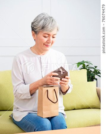 Portrait of an elderly Asian woman receives gift boxes, smiling and looking down while standing in a living room at home. 76074479