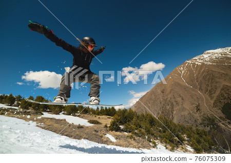 Snowboarder woman jumping from a kicker springboard from the snow on a sunny day in the mountains in a homemade snowboard park Snowboarder woman jumping from a kicker springboard from the snow on a sunny day in the mountains in a homemade snowboard park 76075309