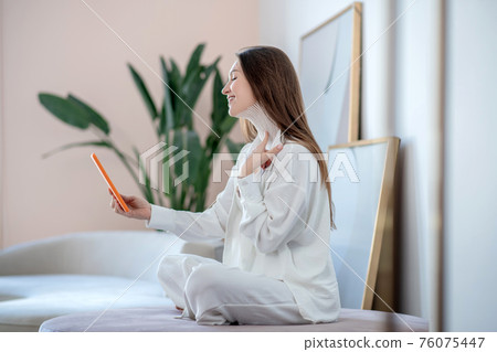 Young woman in white sitting on a soft chair and making a photo of tapes on her neck 76075447