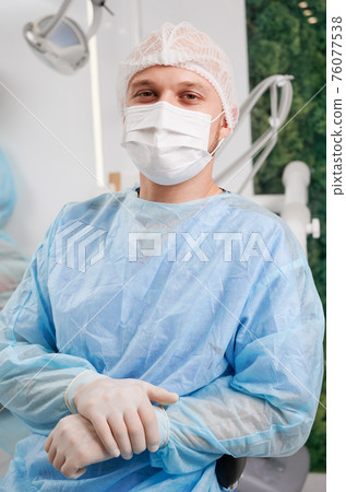 Portrait of young male dentist wearing disposable clothes sitting in modern clinic 76077538