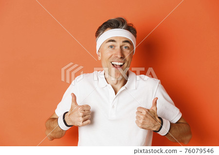 Close-up of cheerful middle-aged male athlete, wearing headband and t-shirt for workout, showing thumbs-up, approve something good, standing over orange background Close-up of cheerful middle-aged male athlete, wearing headband and t-shirt for workout, showing thumbs-up, approve something good, standing over orange background 76079546