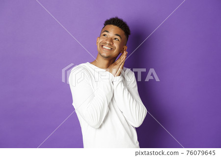 Image of happy african-american man in white sweatshirt, feeling pleased, looking dreamy at upper left corner and smiling, imaging something, standing over purple background 76079645