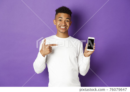 Portrait of handsome african-american man, smiling and pointing finger at smartphone screen, showing application, standing over purple background 76079647