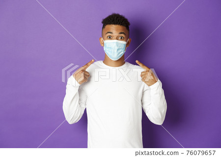 Concept of covid-19, pandemic and social distancing. Image of amused african-american guy, recommend to wear medical mask during coronavirus, standing over purple background Concept of covid-19, pandemic and social distancing. Image of amused african-american guy, recommend to wear medical mask during coronavirus, standing over purple background 76079657