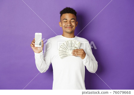 Portrait of pleased african-american man, close eyes and smiling delighted, showing money and smartphone screen, standing over purple background Portrait of pleased african-american man, close eyes and smiling delighted, showing money and smartphone screen, standing over purple background 76079668