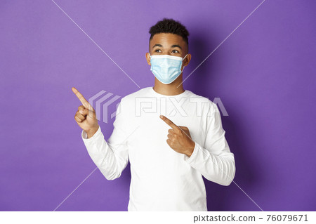 Concept of coronavirus, quarantine and lifestyle. Handsome young african-american man in medical mask, pointing and looking at upper left corner, showing advertisement, posing over purple background 76079671