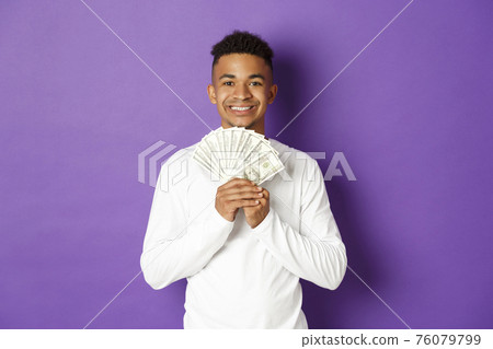 Portrait of handsome african-american man, showing money and smiling, got student loan, standing over purple background 76079799