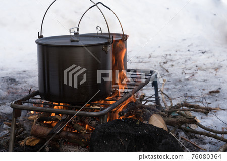 Cooking food on a winter fire. Two beautiful camp pans stand on a grate, under which a fire is made. There is snow all around. Winter tourism. Adventures Cooking food on a winter fire. Two beautiful camp pans stand on a grate, under which a fire is made. There is snow all around. Winter tourism. Adventures 76080364