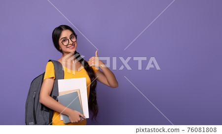 Smiling indian student wearing eyeglasses holding notebooks showing thumbs up Smiling indian student wearing eyeglasses holding notebooks showing thumbs up 76081808