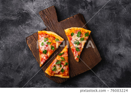 Hot pizza slices with mozzarella cheese, ham, tomato and parsley on brown wooden cutting board, stone concrete background, top view Hot pizza slices with mozzarella cheese, ham, tomato and parsley on brown wooden cutting board, stone concrete background, top view 76083224