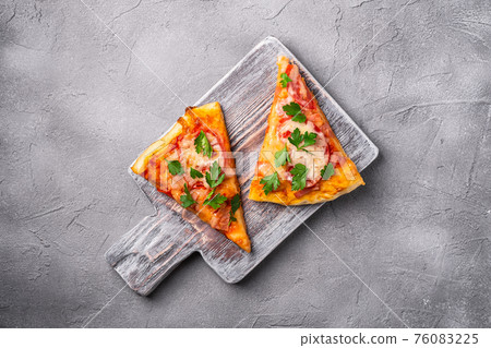 Hot pizza slices with mozzarella cheese, ham, tomato and parsley on wooden cutting board, stone concrete background, top view Hot pizza slices with mozzarella cheese, ham, tomato and parsley on wooden cutting board, stone concrete background, top view 76083225