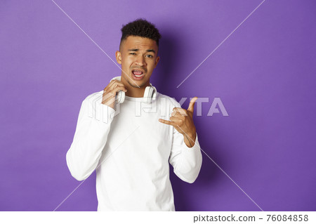 Image of sassy and cool african-american man, take-off headphones and showing hip hop gesture, standing over purple background 76084858