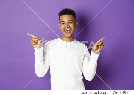 Image of cheerful african-american guy in white sweatshirt, showing two variants, pointing fingers sideways at copy space and smiling, demonstrate products, purple background Image of cheerful african-american guy in white sweatshirt, showing two variants, pointing fingers sideways at copy space and smiling, demonstrate products, purple background 76084935