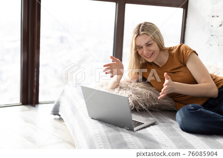 Online video call. Typical adult caucasian woman in casual wear sits on the bed and using laptop at home, selective focus. 76085904
