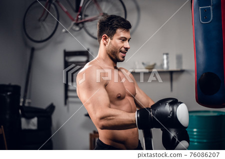 a young athlete with a muscular torso is warming up with a punching bag in his garage. 76086207