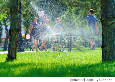 city park on a summer day, green lawns with grass and trees, paths and benches, people walking and children playing, bright sunlight and shadows 76086862