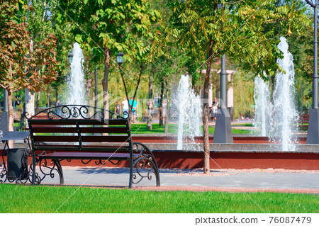 city park on a summer day, green lawns with grass and trees, path, benches and fountain, bright sunlight and shadows 76087479