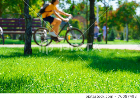 a cyclist rides a bicycle in a city park, summer day, green lawn with grass and trees, bright sunlight and shadows 76087498