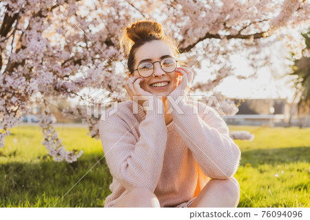 Portrait of young caucasian brunette hipster girl in glasses and light pink clothes sitting on green grass near blooming tree in park. Generation Z girl enjoy spring mood. Selective focus, copy space. 76094096