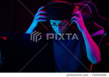 A young esports player sits at a computer during an online game tournament. Neon lighting of the esports arena. A hat and headphones with a headset. 76094806