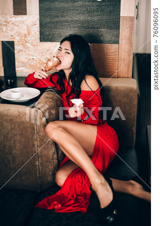 sexy woman in a red dress with black hair eats a croissant. temptation with food. attractive legs. obsession with red. Selective focus, film grain 76096095