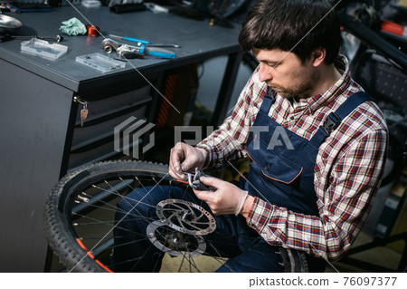 Young caucasian repairman repairing mountain bike wheel in bike shop workshop. Fixing bicycle. Caring for you wheels. man mechanic working in garage. Worker fixing cycle gear. Small business theme 76097377