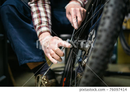 Male mechanic working in bicycle repair shop, mechanic repairing bike using special tool, wearing protective gloves. Young attractive serviceman fixing customer's bicycle wheel at his own workshop 76097414