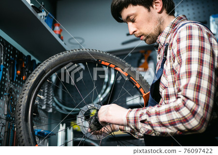 Bicycle mechanic fixing cycle wheel in repair shop. Velocipede service. Repairer male bike shop engineer fix bicycle wheel in workshop. Technician fixes bike parts. Handyman at work in the workplace Bicycle mechanic fixing cycle wheel in repair shop. Velocipede service. Repairer male bike shop engineer fix bicycle wheel in workshop. Technician fixes bike parts. Handyman at work in the workplace 76097422