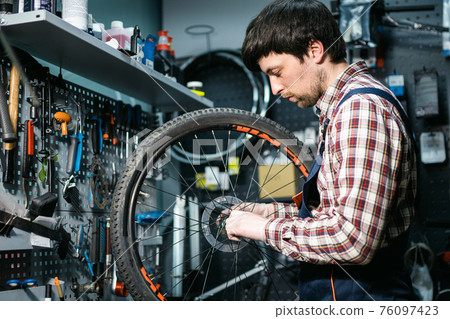 Technical expertise taking care bicycle shop. Handsome young mechanic fixing cycle wheel in workshop. Handsome repairman in workwear serving mountain bicycle. Male engineer adjusting velocipede Technical expertise taking care bicycle shop. Handsome young mechanic fixing cycle wheel in workshop. Handsome repairman in workwear serving mountain bicycle. Male engineer adjusting velocipede 76097423