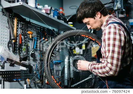 Male mechanic working in bicycle repair shop, mechanic repairing bike using special tool, wearing protective gloves. Young attractive serviceman fixing customer's bicycle wheel at his own workshop 76097424