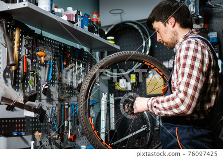 Bike shop mechanic fixing bicycle wheel in workshop. Serviceman repair, maintenance cycle. Velocipede repairing bicycle in bike shop. Bicycle repair service concept. Environmentally friendly vehicles Bike shop mechanic fixing bicycle wheel in workshop. Serviceman repair, maintenance cycle. Velocipede repairing bicycle in bike shop. Bicycle repair service concept. Environmentally friendly vehicles 76097425