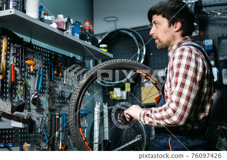 Young caucasian repairman repairing mountain bike wheel in bike shop workshop. Fixing bicycle. Caring for you wheels. man mechanic working in garage. Worker fixing cycle gear. Small business theme Young caucasian repairman repairing mountain bike wheel in bike shop workshop. Fixing bicycle. Caring for you wheels. man mechanic working in garage. Worker fixing cycle gear. Small business theme 76097426