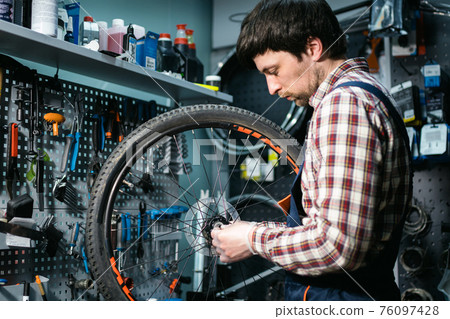 Technical expertise taking care bicycle shop. Handsome young mechanic fixing cycle wheel in workshop. Handsome repairman in workwear serving mountain bicycle. Male engineer adjusting velocipede Technical expertise taking care bicycle shop. Handsome young mechanic fixing cycle wheel in workshop. Handsome repairman in workwear serving mountain bicycle. Male engineer adjusting velocipede 76097428