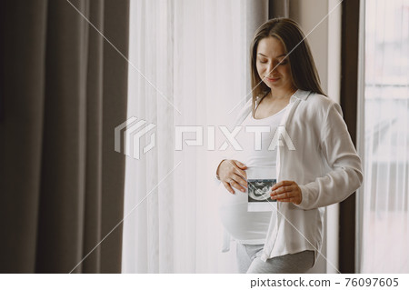 Pregnant woman stands by the window and looks at photo Pregnant woman stands by the window and looks at photo 76097605