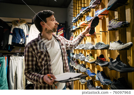 Sporting goods store owner with clipboard checking inventory. Tourist store manager working near showcase with hiking boots makes check list of orders. Salesman exhibit display of walking boots Sporting goods store owner with clipboard checking inventory. Tourist store manager working near showcase with hiking boots makes check list of orders. Salesman exhibit display of walking boots 76097968
